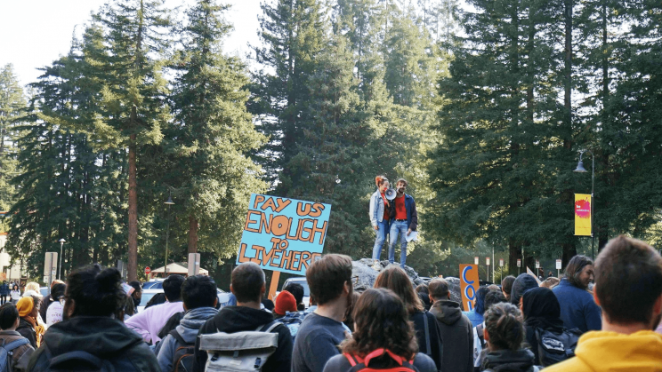 Dozens of student protesters are gathered around two organizers who are standing on a rock with a megaphone. One protester holds a sign "Pay Us Enough To Live Here" with orange letters on a light-blue field. In the background of the image are tall coniferous trees particular to California and the West Coast.