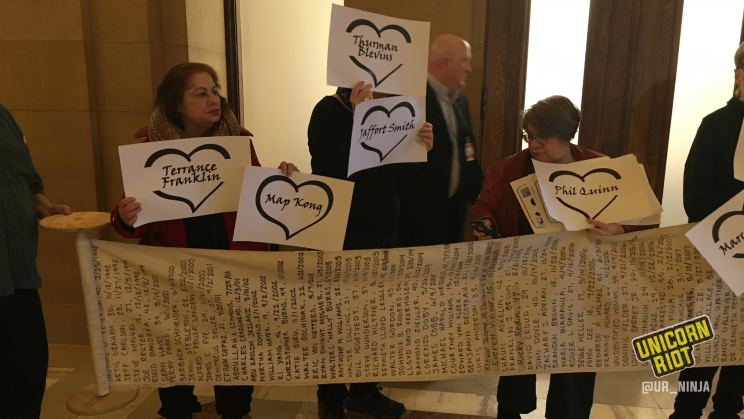 Several community members are holding up individual signs each featuring the name of a person who had been killed by the police. The community members are standing behind a long scroll of paper unrolled horizontally that lists hundreds of names of people killed by police.