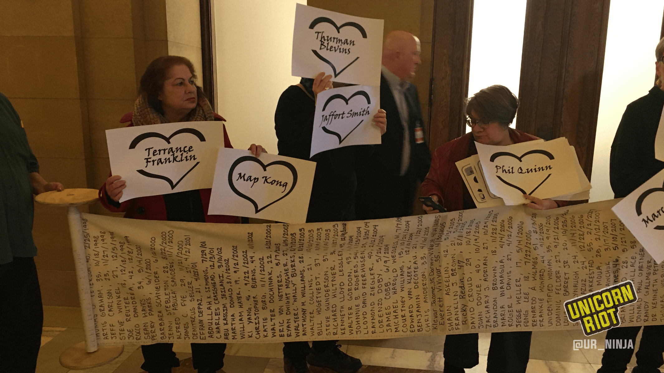 Several community members are holding up individual signs each featuring the name of a person who had been killed by the police. The community members are standing behind a long scroll of paper unrolled horizontally that lists hundreds of names of people killed by police.