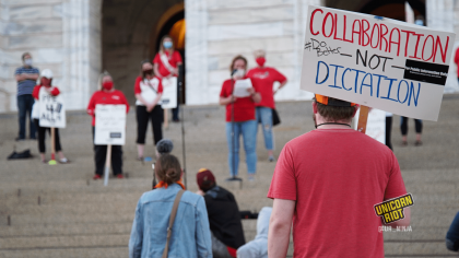 image: a protester holds a sign, "Collaboration - Not - Dictation. #DoBetter" They are wearing a red t-shirt and are wearing an orange-and-black baseball cap. In the background are protesters standing ~6' apart on the steps of the Capitol building in Saint Paul.