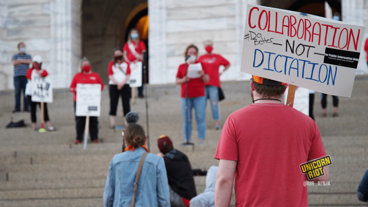 image: a protester holds a sign, "Collaboration - Not - Dictation. #DoBetter" They are wearing a red t-shirt and are wearing an orange-and-black baseball cap. In the background are protesters standing ~6' apart on the steps of the Capitol building in Saint Paul.