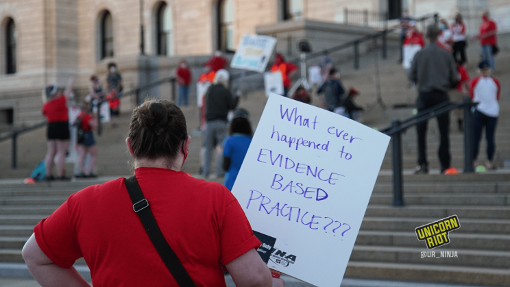 image: a protester stands facing away from the camera, holding a sign, "What ever happened to EVIDENCE BASED PRACTICE???" They are wearing a red t-shirt and a black bag with a black strap slung diagonally over their shoulders. In the background are protesters standing ~6' apart on the steps of the Capitol building in Saint Paul.