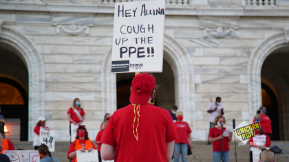 image: a protester holds a sign, "Hey Allina, Cough Up The PPE." The person is wearing a backwards red baseball cap and a red shirt. In the background are protesters standing ~6' apart on the steps of the Capitol building in Saint Paul.