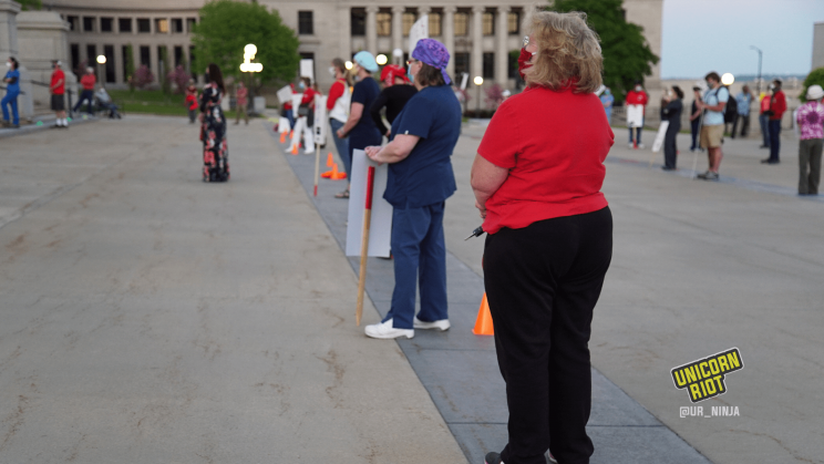 Image: medical professionals and supporters stand in a row, each 6 feet or 2 meters apart from the next, spaced out by orange cones placed on the ground. Every protester is wearing a cloth mask covering their nose and mouth, as this photo was taken during the pandemic of 2020.