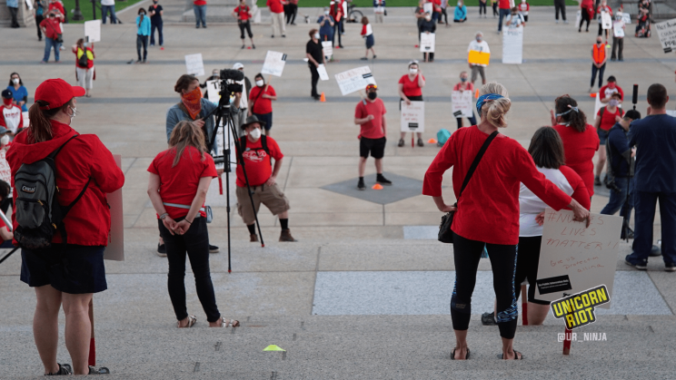 image: Looking down from in front of the entrance to the Minnesota Capitol building, protesters (all of whom are wearing masks, most of whom are wearing red) are spaced six feet apart from each other (two meters) so as to maintain safe physical distance while protesting.