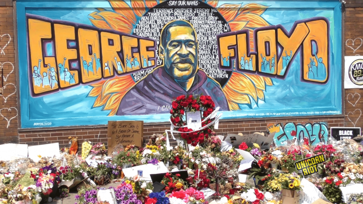 image: George Floyd mural at the vigil site at 38th & Chicago, as seen by day time. In the center is an image of George Floyd in front of a large sunflower, in the center of which is painted the names of others lost to police terror, including Breonna Taylor. The sunflower and George Floyd's name are yellow and orange, and the background of the mural is blue. In front of the mural on the ground are hundreds of flowers and #JusticeForGeorgeFloyd signs. A heart-shaped wreath of red roses has been placed in front of the mural as a memorial.