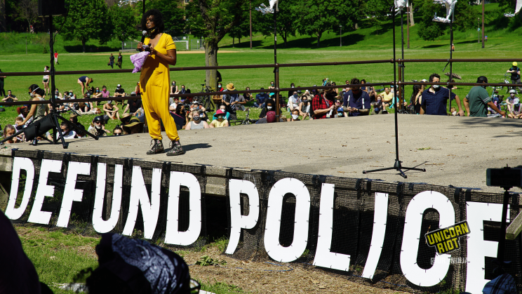 image: Black Visions organizer Miski Noor stands on top of the stage addressing the gathered crowd, who is seated in front of her and to her right behind her. She is wearing a saffron-colored short-sleeved one-piece. Across the stage below her are the words DEFUND POLICE in large capital white letters on a black semi-opaque backround.