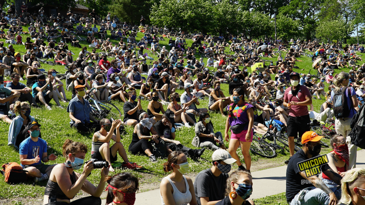 Image: hundreds of people are seated on the hill at Powderhorn Park that slopes down to the sidewalk that runs the perimeter of the lake. Everyone is wearing fabric face masks.