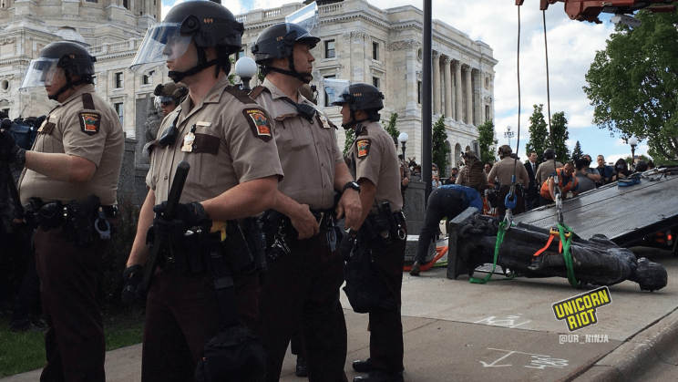 image: state troopers with weapons stand between community members and the statue of Christopher Columbus which was toppled from its pedestal on the grounds of the Minnesota State Capitol on June 10, 2020. The statue is face-down on the ground. Straps are wrapped around the statue – it is about to be hoisted into the air and then placed on a flat-bed truck for removal.
