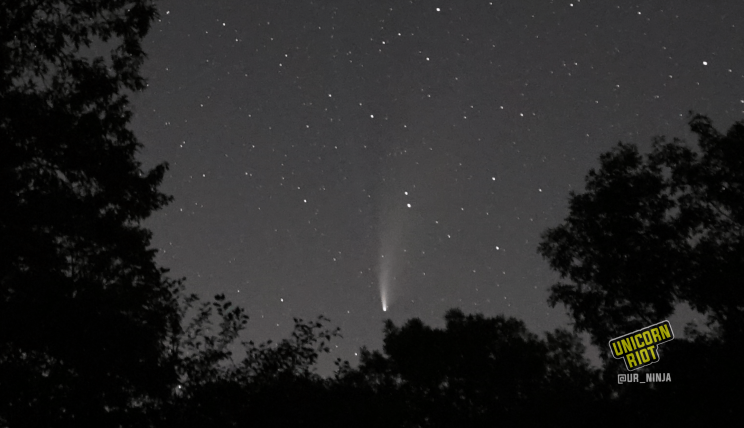 image: ten-second long exposure shot of the night sky facing North, in the northern hemisphere at midsummer. Both the dust tail as well as the ion gas tail of comet NEOWISE are visible, pointing upwards perpendicular to the horizon.