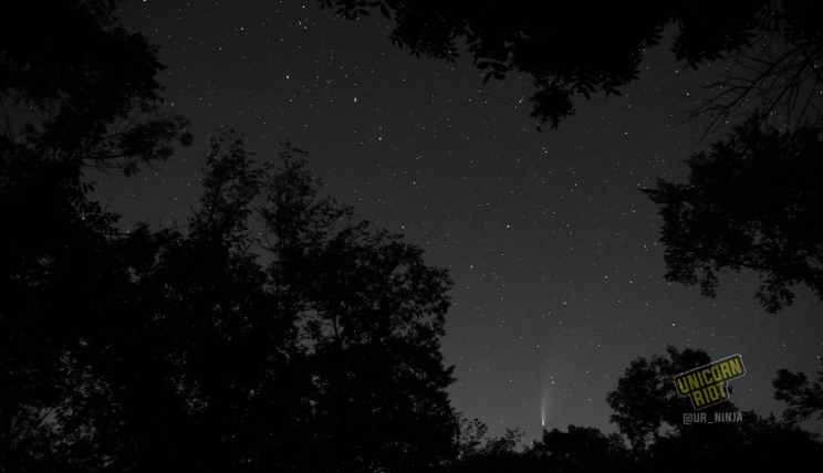 image: ten-second long exposure shot of the night sky facing North, in the northern hemisphere at midsummer. Comet NEOWISE is visible near the tree line at the bottom of the image. The seven familiar stars forming the familiar handle/ladle of "the big dipper" are visible in the center of the image. A slight glow is visible on the bottom of the image from a nearby city.