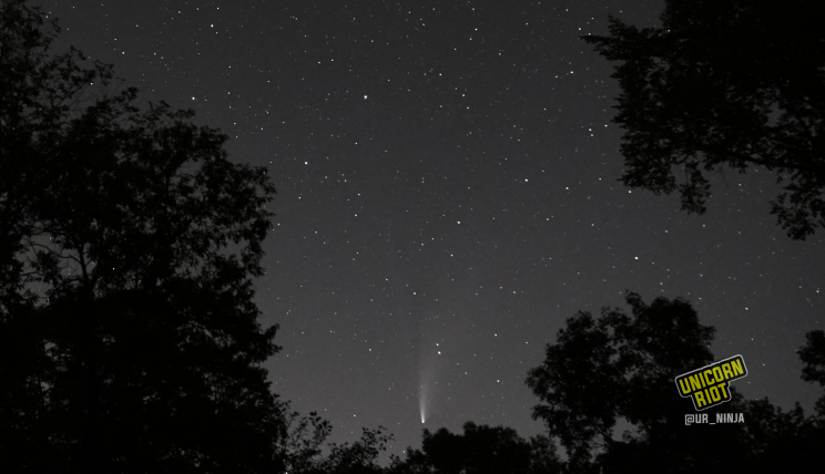 image: ten-second long exposure shot of the night sky facing North, in the northern hemisphere at midsummer. Both the dust tail as well as the ion gas tail of comet NEOWISE are visible, pointing upwards perpendicular to the horizon.