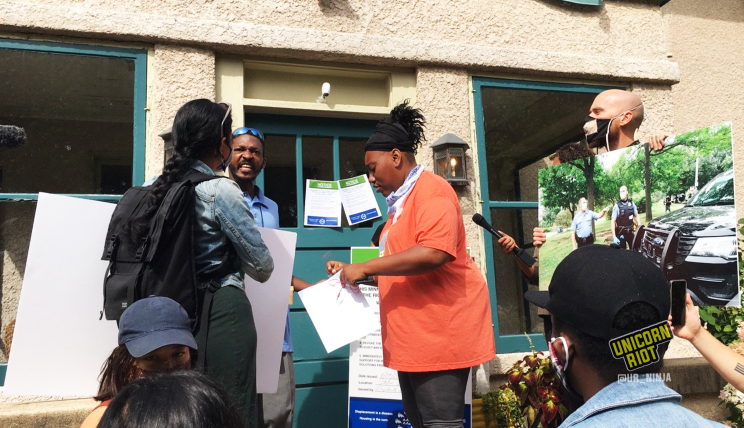 image: a Black woman in an orange shirt is adding tape to a piece of paper, a list of demands for the Minneapolis Parks & Recreation Board Superintendent, Al Bangoura. On the woman's left is a Lakota man with chest-length black hair, as well as a media-maker with their back turned. On the right is a member of Minneapolis Parks & Power; he is wearing a mask and holding a blown-up photograph from when Minneapolis Police assisted in the violent clearing of the sanctuary encampment at Powderhorn Park on Friday, August 14, 2020.