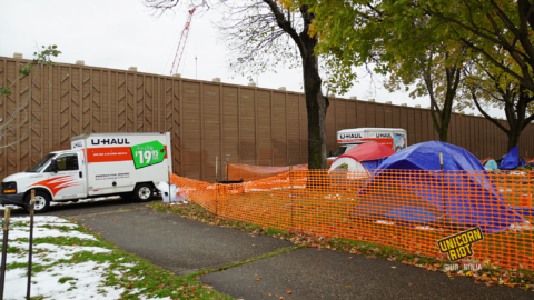 image: the perimeter of a sanctuary tent camp area is surrounded by orange plastic construction fencing; the fence cuts diagonally across the image from bottom right, to the left of the image. On the sidewalk outside of the fencing, two U-Haul trucks are parked; residents' belongings are inside, being moved from MLK Park to another location. A gray tent with a blue tarp is propped up beneath and to the left behind the trunk of a maple tree. To the right of the tree is a waste bin that says City of Minneapolis on its side. Partially-melted snow is visible on the ground.