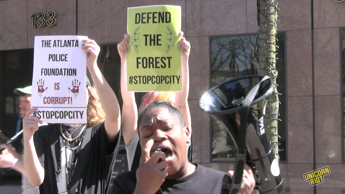 Reverend Keyanna Jones, a Black woman with her hair tied back, speaks into a black megaphone as two young white protesters stand behind her holding up signs that say "The Atlanta Police Foundation is Corrupt" and "Defend the Forest." The signs block the two protesters' faces. This photo shows these three at a protest outside the downtown Atlanta headquarters of 'Cop City' sponsor corporation Georgia Pacific's offices at 133 Peachtree St. A larger crowd stands out of frame behind the camera.