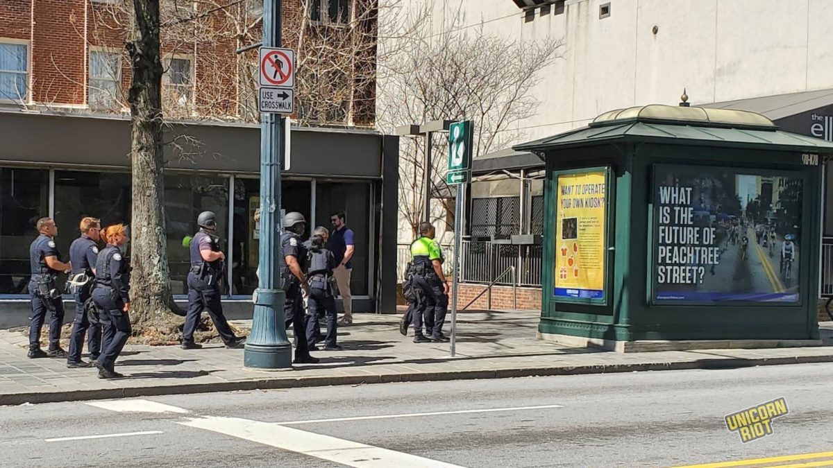 A formation of 8 Atlanta Police officers marches on the sidewalk during a small downtown Stop Cop City protest march on March 7, 2023. Two of the 8 officers wear reinforced black helmets and at least one of them is carrying an assault rifle. The cluster of cops is about to pass a newspaper vending kiosk with a large advertisement poster on it reading "What is the future of Peachtree Street?"

Out of frame, the protest march is on the parallel sidewalk across the street.  