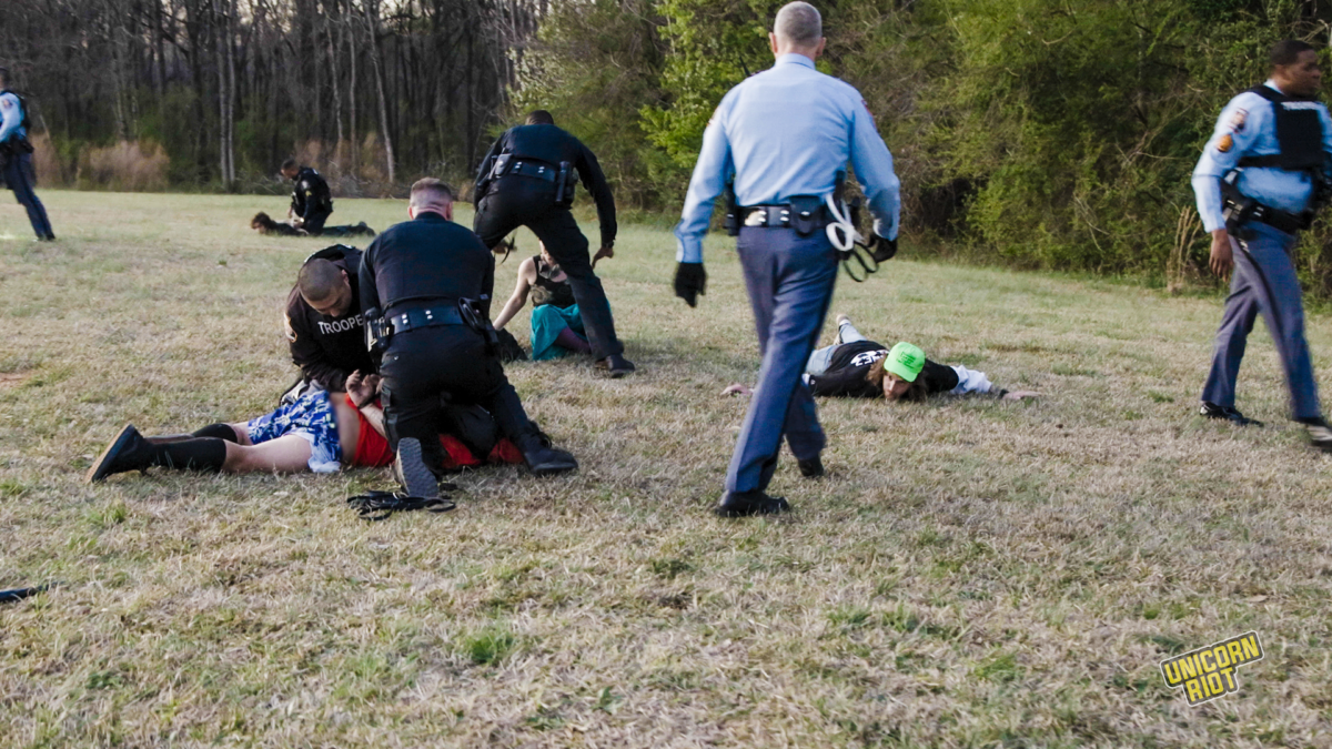 Georgia State Troopers making arrests after storming the Stop Cop City music festival in the "RC Field" clearing in the South River Forest near Atlanta on March 5, 2023. On the left, one arrestee is pinned down my two officers in dark blue kneeling on them. In the middle-left of the shot, a person sitting down can be seen sitting and about to be arrested. On the right center part of the image, a man in a neon green National Lawyers Guild legal observer hat is lying down on the ground with his hands out to the sides - presumably having done so on orders by an arresting officer. A Georgia State Patrol higher-up is seen striding towards the prone NLG observer with black leather gloves on his hands; other officers in frame do not appear to be wearing gloves.