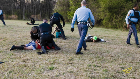 Georgia State Troopers making arrests after storming the Stop Cop City music festival in the "RC Field" clearing in the South River Forest near Atlanta on March 5, 2023. On the left, one arrestee is pinned down my two officers in dark blue kneeling on them. In the middle-left of the shot, a person sitting down can be seen sitting and about to be arrested. On the right center part of the image, a man in a neon green National Lawyers Guild legal observer hat is lying down on the ground with his hands out to the sides - presumably having done so on orders by an arresting officer. A Georgia State Patrol higher-up is seen striding towards the prone NLG observer with black leather gloves on his hands; other officers in frame do not appear to be wearing gloves.