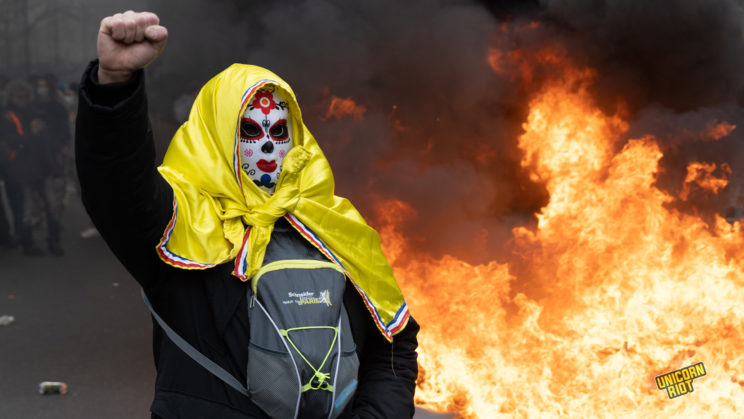 person with a mask on and yellow flag around their head and backpack in the front of their body stand in front of fire posing with their fist in the air during protests in Paris on March 28, 2023