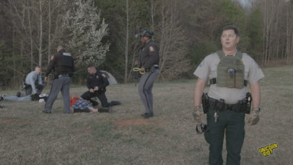 A white Georgia Department of Natural Resources game warden wears a small bulletproof vest with his belly protruding underneath looks to the side with his mouth agape, his face featuring a slightly phased and aloof look. Behind him and to the viewer's left, two people are face down in the process of being arrested by 4 Goergia state troopers - three troopers are white, one is black. They are wearing brown, black or light blue uniform shirts with pin striped gray pants. Most of them have plastic zip tie 'flex-cuffs' in hand as part of the arrest process taking place in the RC Field area of the South River Forest during the 'Stop Cop City' music festival on March 5, 2023. Trees can be seen in the forest behind the arrest scene; all is bathed in a pale faintly orange light as the sun begins to set.