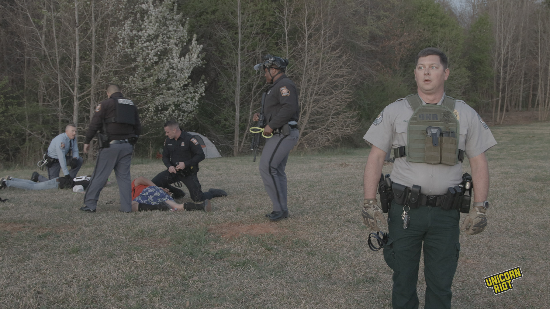 A white Georgia Department of Natural Resources game warden wears a small bulletproof vest with his belly protruding underneath looks to the side with his mouth agape, his face featuring a slightly phased and aloof look. Behind him and to the viewer's left, two people are face down in the process of being arrested by 4 Goergia state troopers - three troopers are white, one is black. They are wearing brown, black or light blue uniform shirts with pin striped gray pants. Most of them have plastic zip tie 'flex-cuffs' in hand as part of the arrest process taking place in the RC Field area of the South River Forest during the 'Stop Cop City' music festival on March 5, 2023. Trees can be seen in the forest behind the arrest scene; all is bathed in a pale faintly orange light as the sun begins to set.