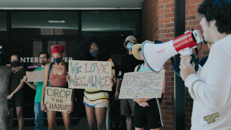 A protester in a white shirt stands on the right edge of the frame holding a megaphone addressing the crowd standing in front of the entrance to the DeKalb County building - signs held by protesters seen in frame read (from right to left): "Abolition is Creative" - "We Love Weelaunee" - "Drop the Charges" and "No Cops No Prisons"