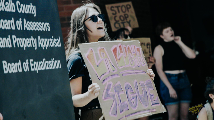 A black monolith with white font reading "DeKalb County Board of Assessors and Property Appraisal... Board of Equalization" is seen to the left of a chanting protester with long hair and sunglasses holding a sign that reads "La Lucha Sigue"