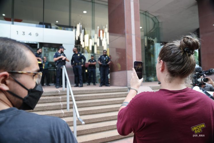 One protester wearing a black KN95 mask looks to the right as another holds up their cell phone to photograph a dozen or so Atlanta Police officers standing in front of the entrance to the building at 1394 W Peachtree Street that includes the offices of Cadence Bank