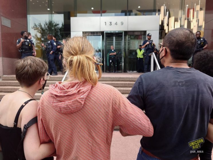 3 protesters backs and heads seen from behind as they face the 1349 W Peachtree Street building housing the offices of Cadence Bank. Most of a dozen Atlanta Police officers can be seen (out of focus) standing in front of the glass floor-to-ceiling t building entrance
