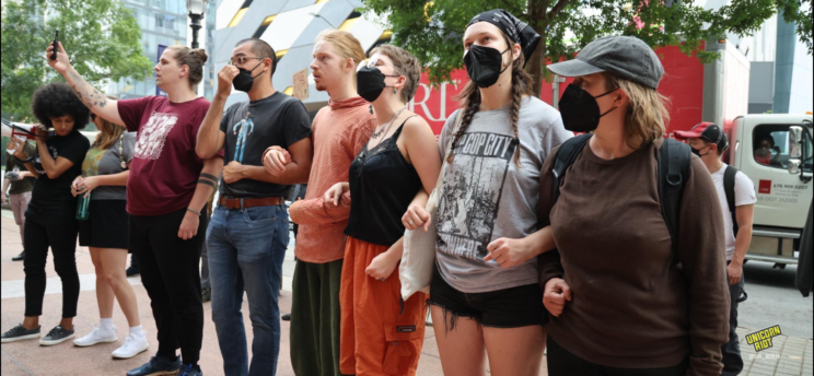 A line of 6 protesters join arms while standing on the sidewalk in front of Cadence Bank - most are wearing black KN95 masks