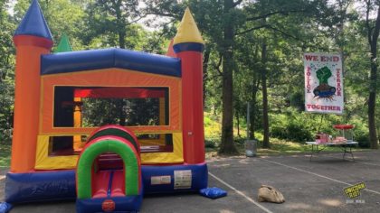 A colorful bouncy castle house with pointy cone tips in its four corner columns sits on the basketball court at Brownwood Park in southeast Atlanta. To its right snacks and a popcorn machine sit on a folding table. Behind the snack table a banner hangs from trees reading “We End Police Terror Together” with a painted image of a clenched green fist emerging from an overturned and smashed police car - brown roots feeding into the green fist can be seen growing into the ground through the smashed windshield of the upside down cop car.
