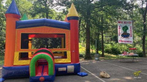 A colorful bouncy castle house with pointy cone tips in its four corner columns sits on the basketball court at Brownwood Park in southeast Atlanta. To its right snacks and a popcorn machine sit on a folding table. Behind the snack table a banner hangs from trees reading “We End Police Terror Together” with a painted image of a clenched green fist emerging from an overturned and smashed police car - brown roots feeding into the green fist can be seen growing into the ground through the smashed windshield of the upside down cop car.