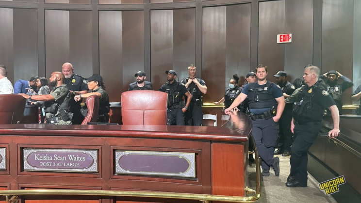 13 Atlanta Police officers stand behind the empty seat in Atlanta City Council chambers labeled" Keisha Sean Waites Post 3 At Large. Lt. Neil Welch leans against a waist-high wall gazing into the distance with sweat on his face and his cell phone in his right hand. To his left a younger officer with a lot of hair gel in his hair stares menacingly directly into the camera.