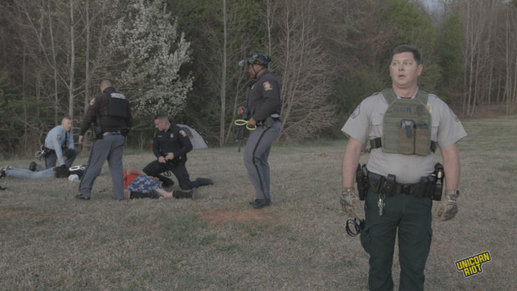 A white Georgia Department of Natural Resources game warden wears a small bulletproof vest with his belly protruding underneath looks to the side with his mouth agape, his face featuring a slightly phased and aloof look. Behind him and to the viewer's left, two people are face down in the process of being arrested by 4 Goergia state troopers - three troopers are white, one is black. They are wearing brown, black or light blue uniform shirts with pin striped gray pants. Most of them have plastic zip tie 'flex-cuffs' in hand as part of the arrest process taking place in the RC Field area of the South River Forest during the 'Stop Cop City' music festival on March 5, 2023. Trees can be seen in the forest behind the arrest scene; all is bathed in a pale faintly orange light as the sun begins to set.
