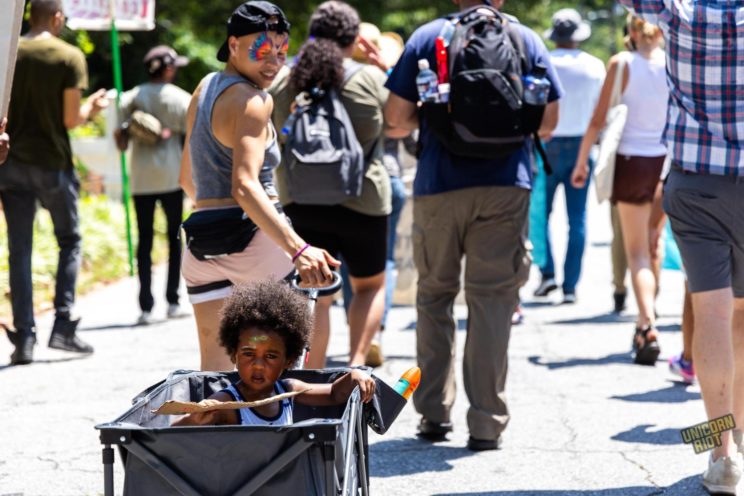 A small Black child with an Afro and red and gfeeen face paint on their forehead is pulled in a wagon by an adult with a Fanny pack and a bright neon butterfly design affixed around their eyes with face paint - they are situated amongst the larger protest crowd, the photo is taken facing the backs of the marchers but the child is facing backwards towards the camera