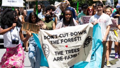 An activist speaking into a megaphone can be seen amidst a partial shot of a marching crowd - a banner being held in the front of the march reads "Don't Cut Down the Forest! The trees are family."