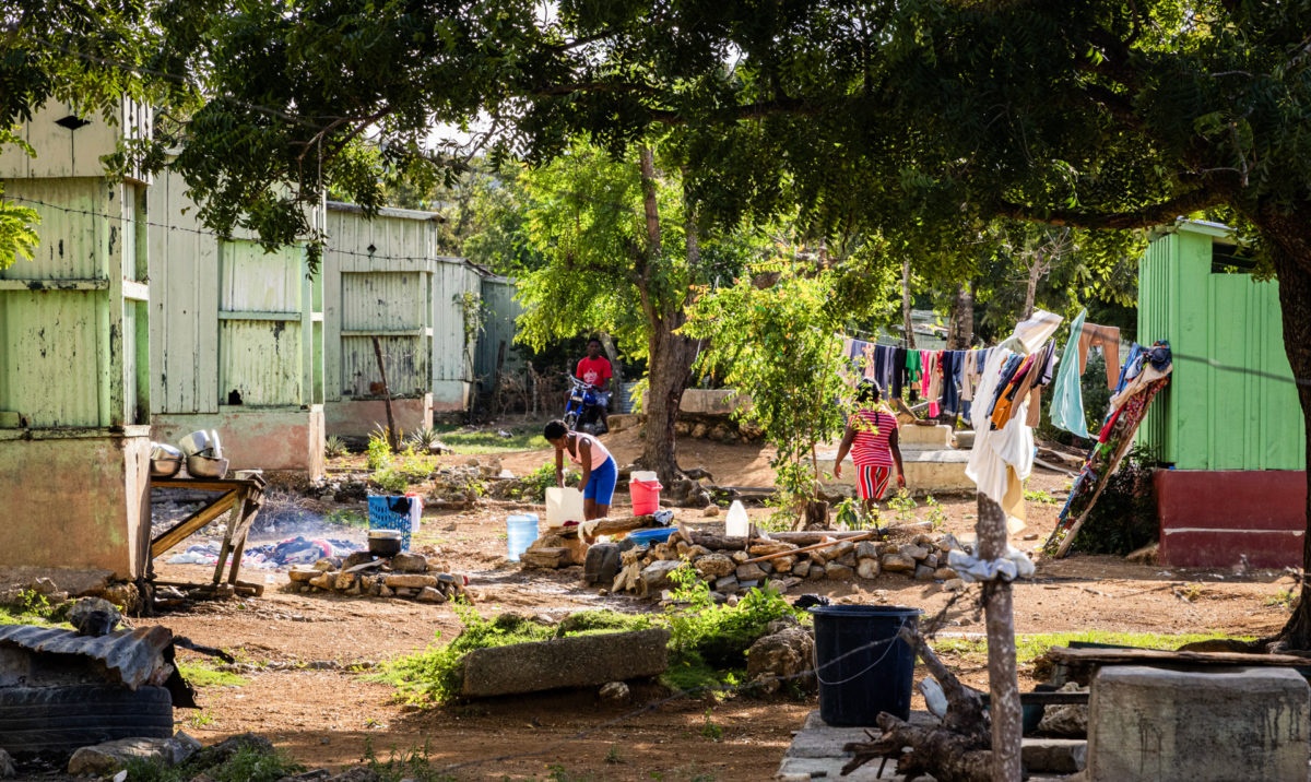 Surrounded by green wooden houses on a cement base, two women hand wash their families' clothes using refillable water jugs then hang them up to dry on a clothesline. A man is seated on a small motorbike behind them.