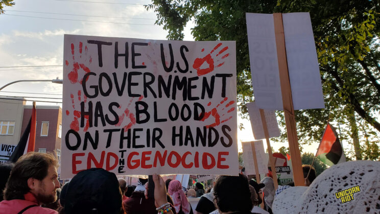 A protest sign features red handprints on a white background alongside black and red text that reads "The U.S. Government Has Blood On Their Hands - End The Genocide" - various protesters are seen marching from behind in a crowd around the sign; two Palestinian flags are slightly visible on either side of the frame