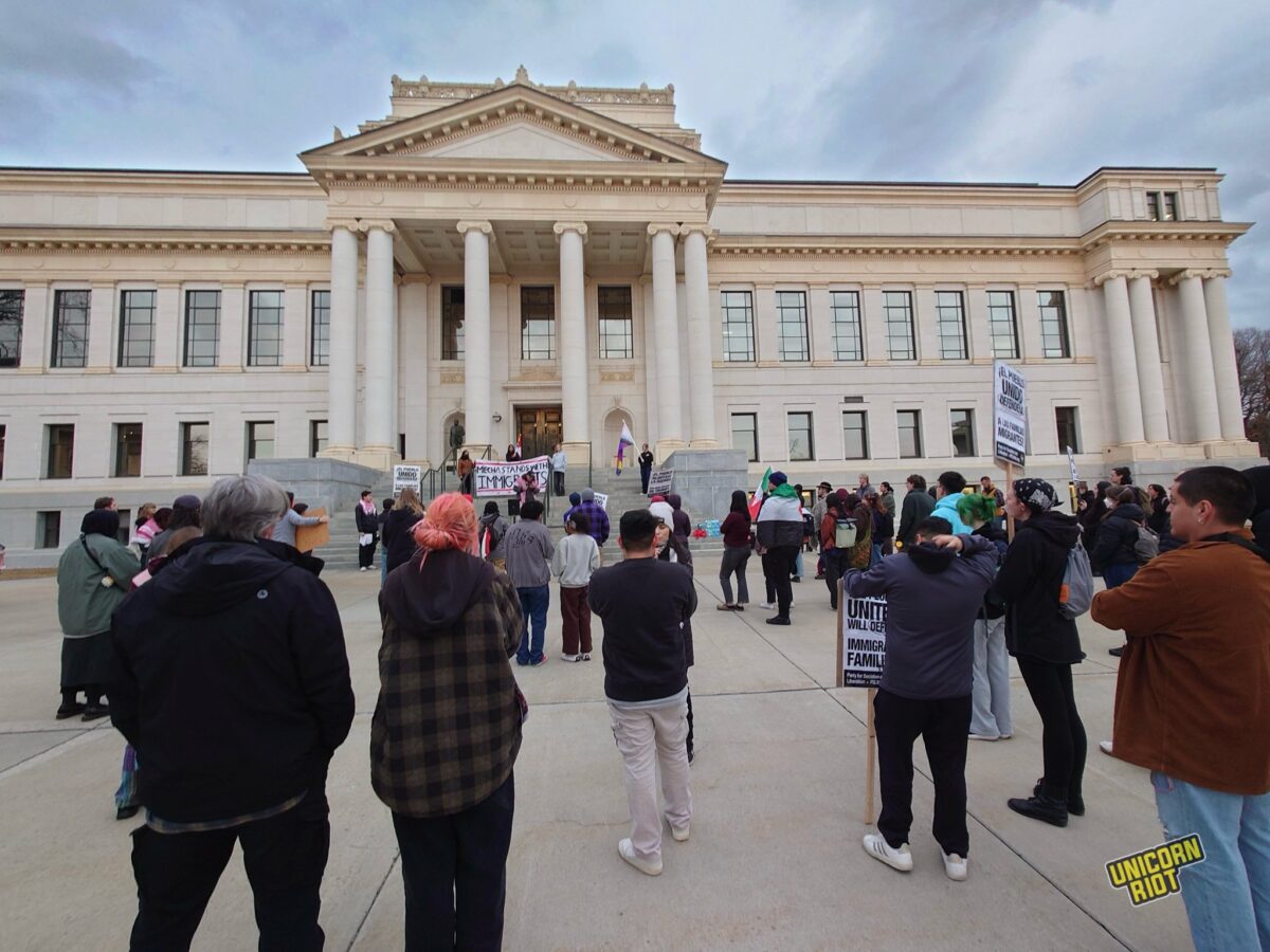 A mixed group of protesters scattered across the concrete plaza in front of a large classical hall building by at the University of Utah seen from behind - in the distance a protest banner can be seen on the building’s steps
