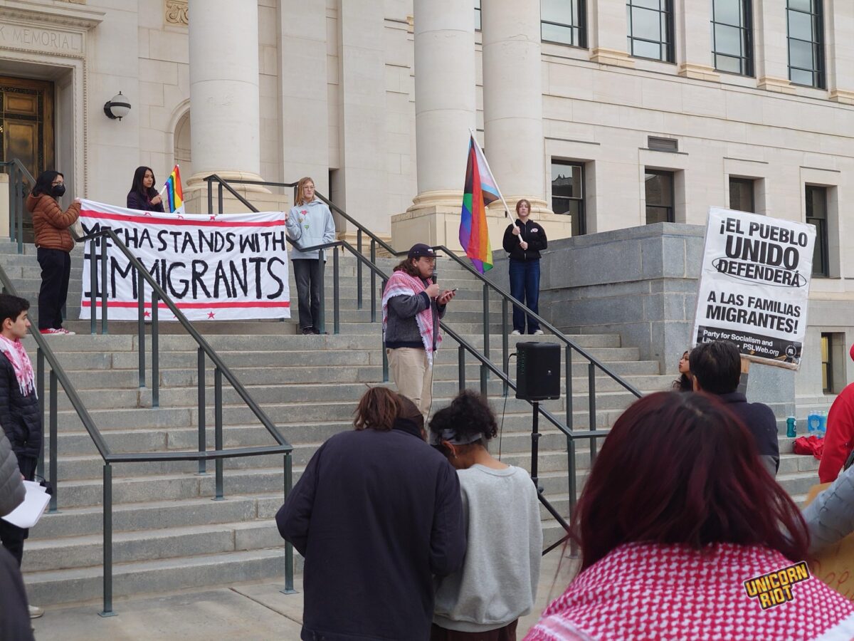 Protesters on the concrete steps of a University of Utah building with large white columns hold a banner that appears to say “MECHA STANDS WITH IMMIGRANTS” - a speaker wearing a red kaffiyeh scarf addresses the crowd while reading off their phone - an identical scarf is seen on a protester close to the camera with their back facing the lens - a visible black and white protest sign in the crowd reads “EL PUEBLO UNIDO DEFENDERA - A LAS FAMILIAS MIGRANTES”