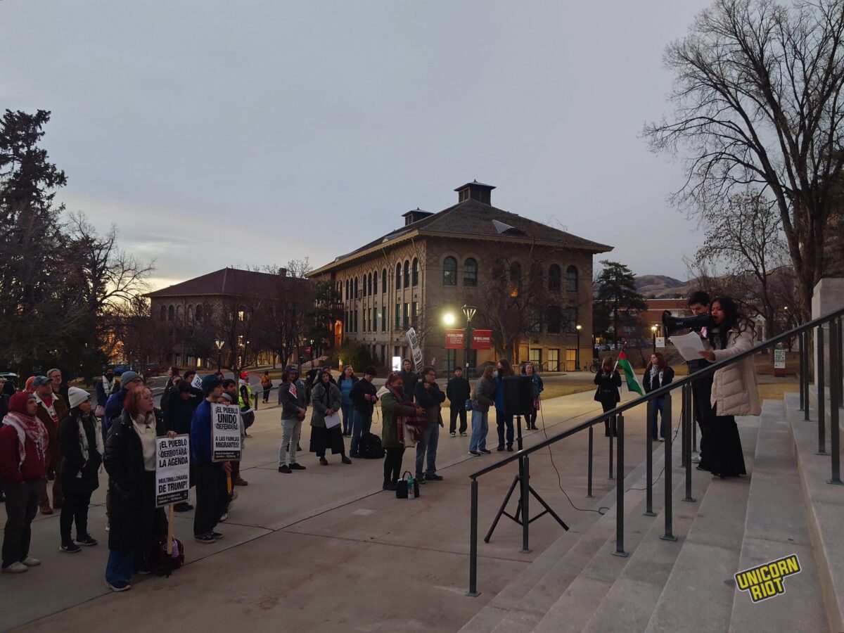 A cluster of protesters, some holding signs, gather at the base of the aforementioned U of U campus building’s large steps as a speaker reading off a piece of paper addresses them with a bullhorn
